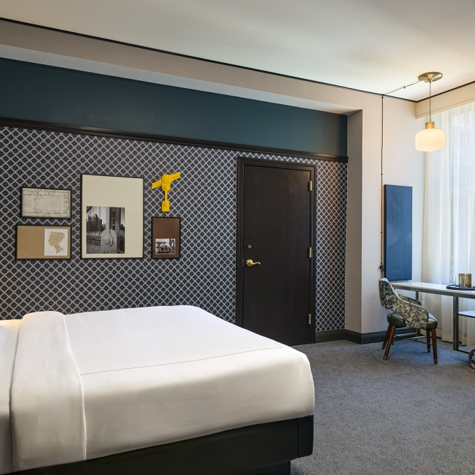 A well-appointed hotel room displays a neatly made bed, geometric patterned walls with framed art, a dark door, and a desk by a window.