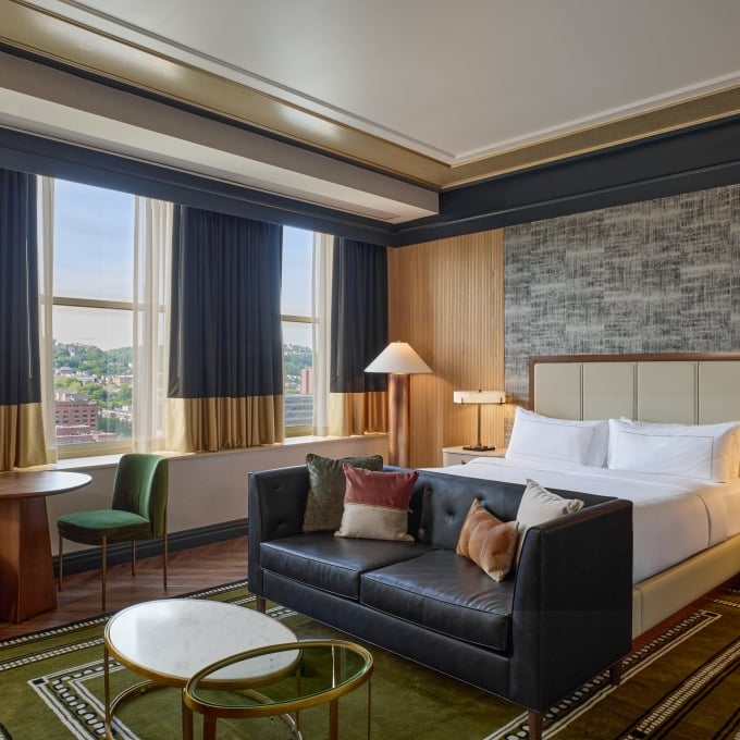 Luxury hotel room with a king bed, a sleek dark leather sofa, a round wooden dining table, and large window curtains.