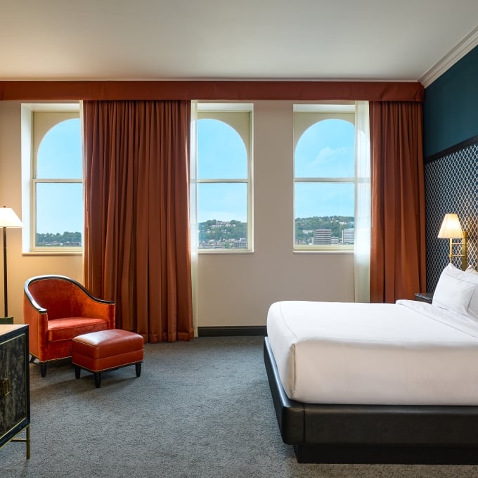 Luxury hotel bedroom with blue geometric wallpaper, dark leather bed, orange leather armchair, and a view of city windows.