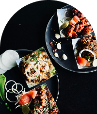 Several plates of various Indian-style dishes are artfully arranged and garnished with onions, tomatoes, and garlic on a dark, textured surface.