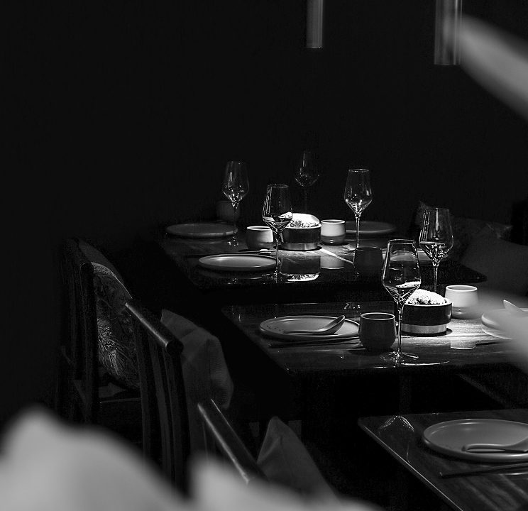 Tables are set with plates, wine glasses, and bowls, waiting for diners in a dimly lit, high-contrast restaurant scene, rendered in black and white.