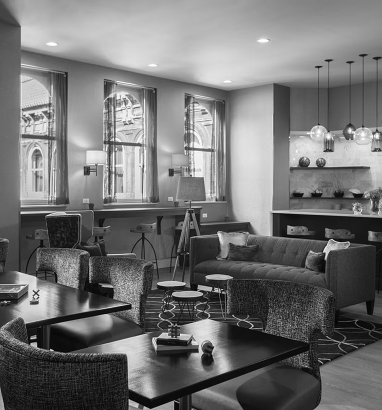 A chic lounge area furnished for comfort, featuring various dark tables, modern seating, and pendant lights. Large windows reveal an ornate building outside in this black and white interior.