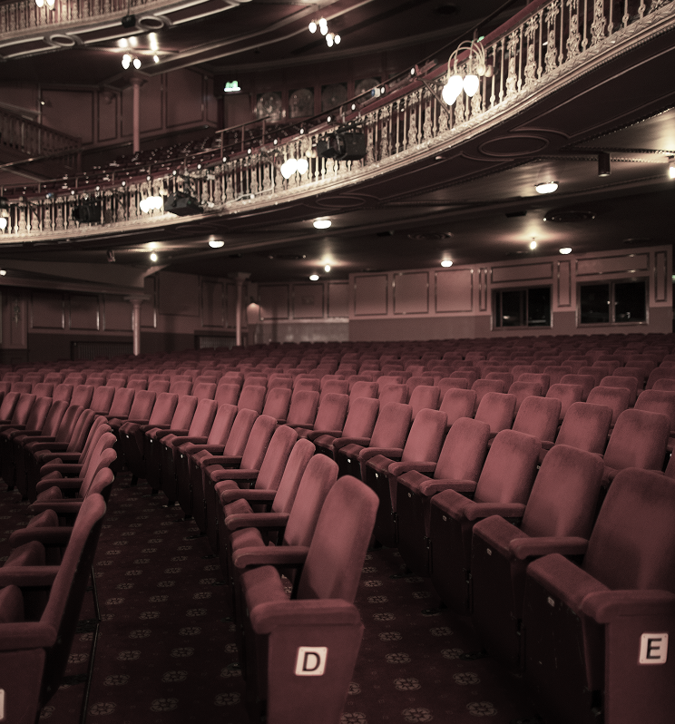 Rows of empty, maroon velvet theater seats are arranged facing forward, with an ornate balcony overhead, in a dimly lit auditorium. Seat labels 'D' and 'E' are visible.