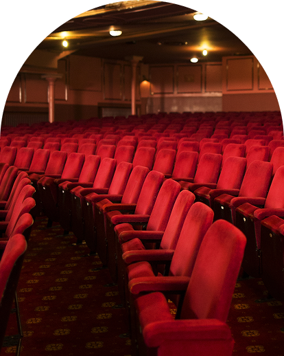 Red velvet theater seats are arranged in curving rows on a patterned red carpet, filling a dimly lit, ornate auditorium.