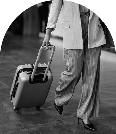 A person in striped pants and a jacket walks, pulling a small silver rolling suitcase across a tiled floor indoors.