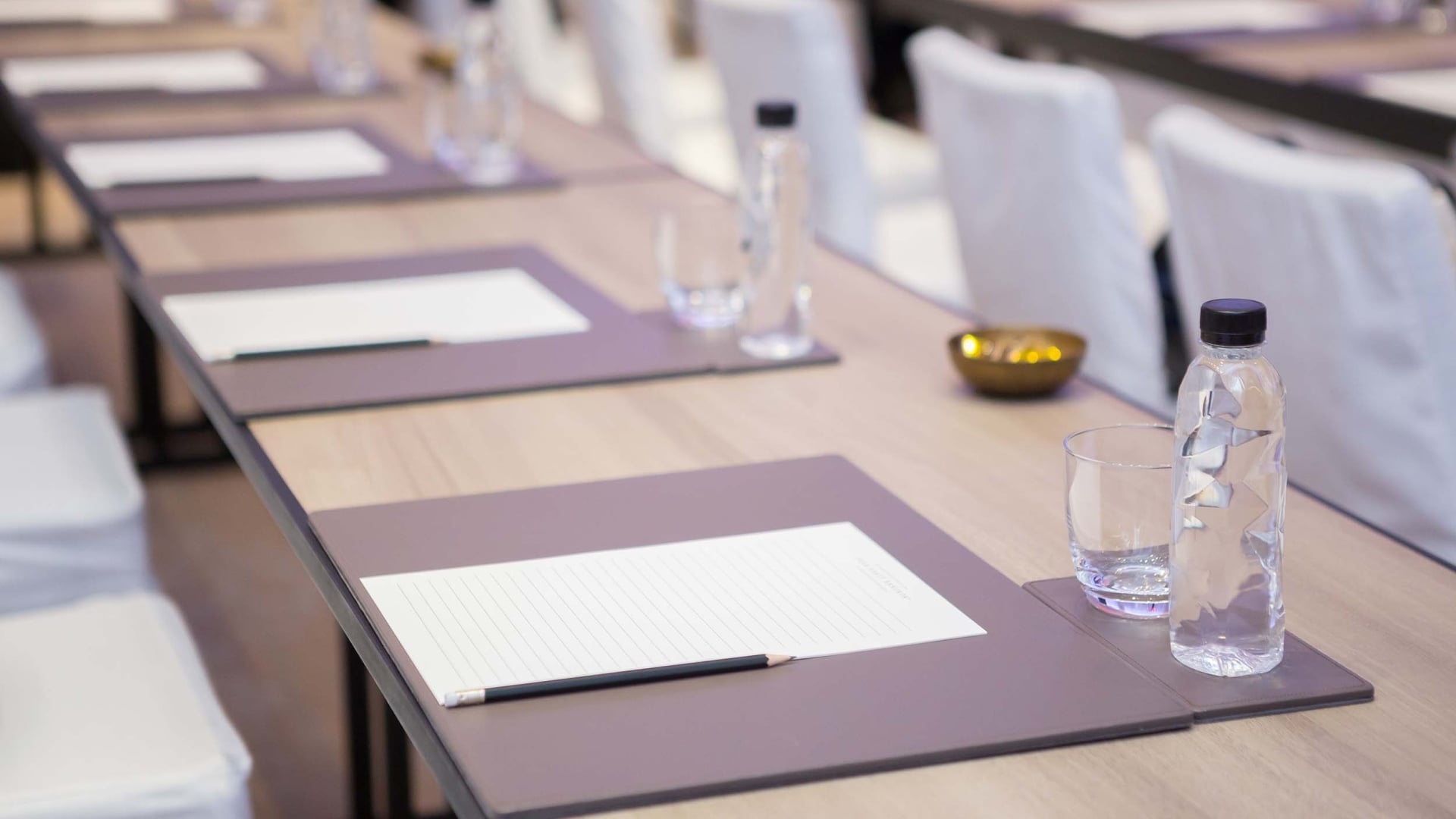 Conference table set with individual place settings, each having a notepad (MANDARIN ORIENTAL BANGKOK), pencil, bottled water, and glass, ready for a meeting.