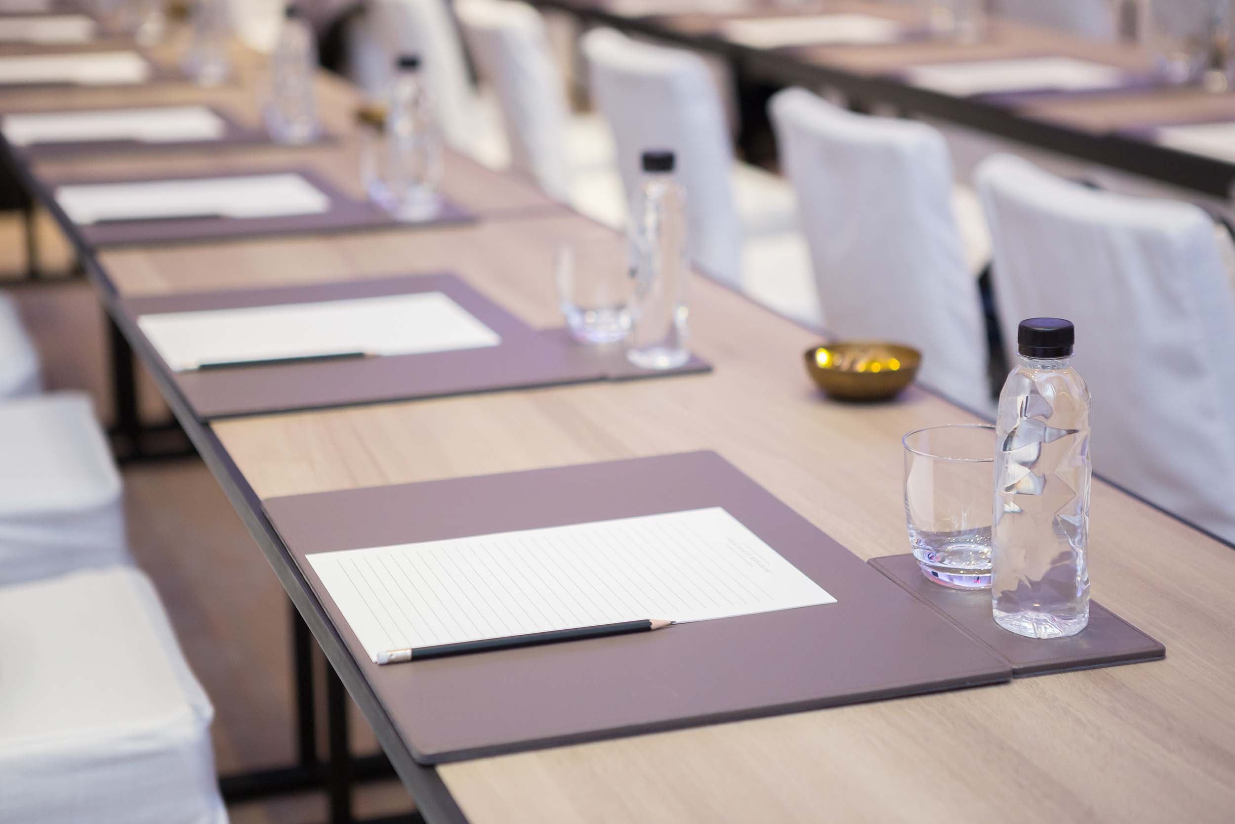 Conference table set with individual place settings, each having a notepad (MANDARIN ORIENTAL BANGKOK), pencil, bottled water, and glass, ready for a meeting.
