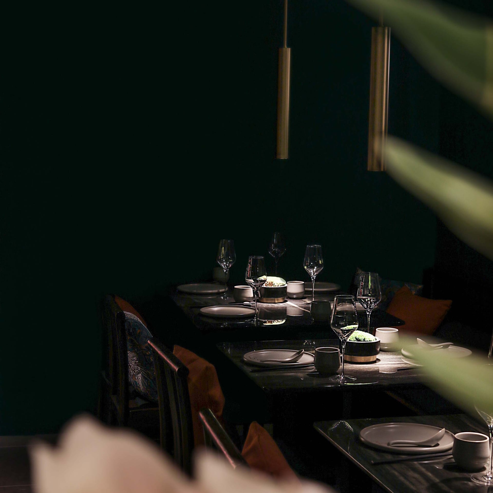 Multiple dining tables, set with elegant dinnerware and wine glasses, are arranged in a dimly lit, dark green restaurant interior, partially obscured by blurred foreground plants.