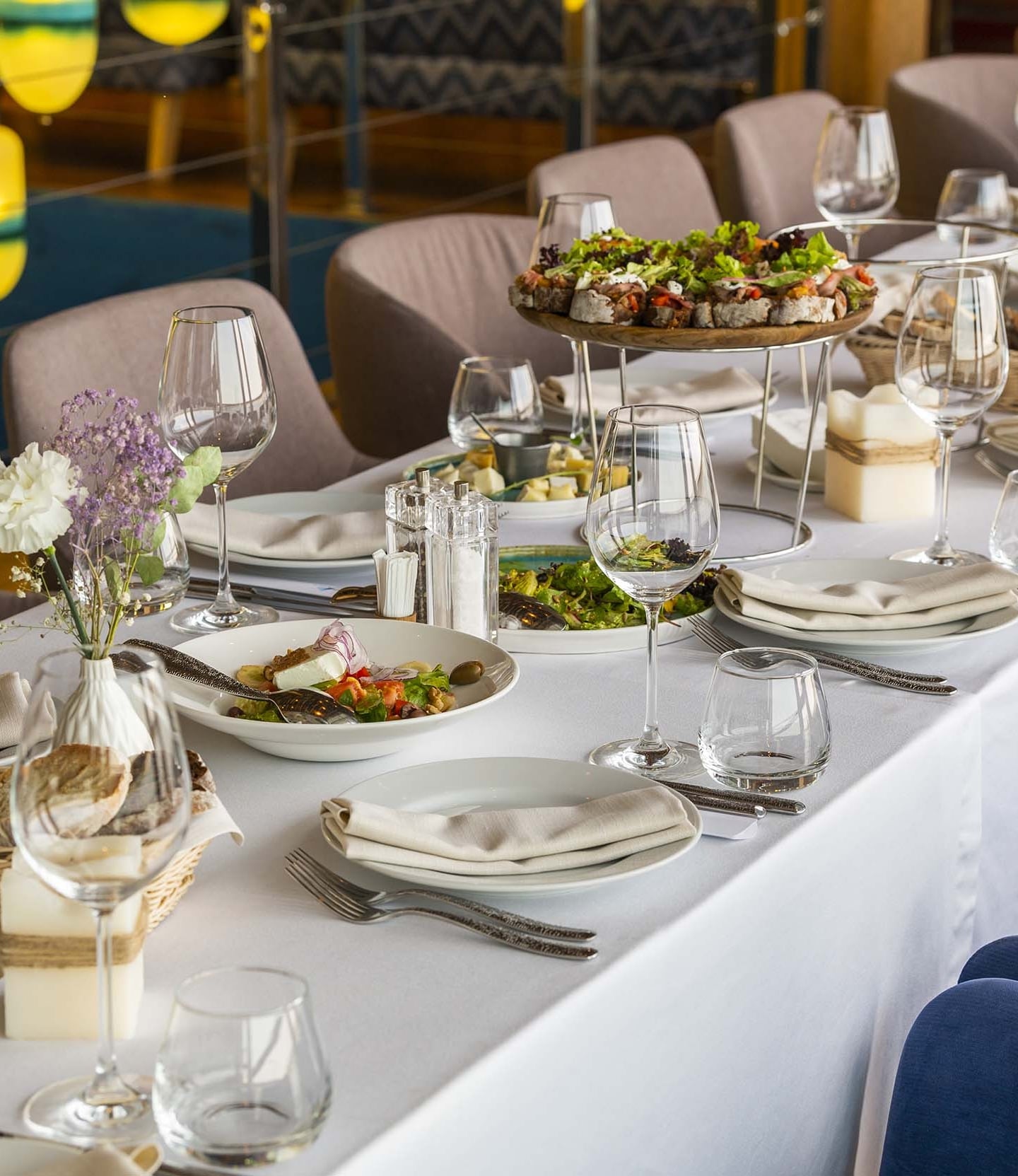 A long dining table is lavishly set with various dishes, glasses, and food, including salads and canapés, inside a well-lit restaurant with colorful yellow and blue accents.