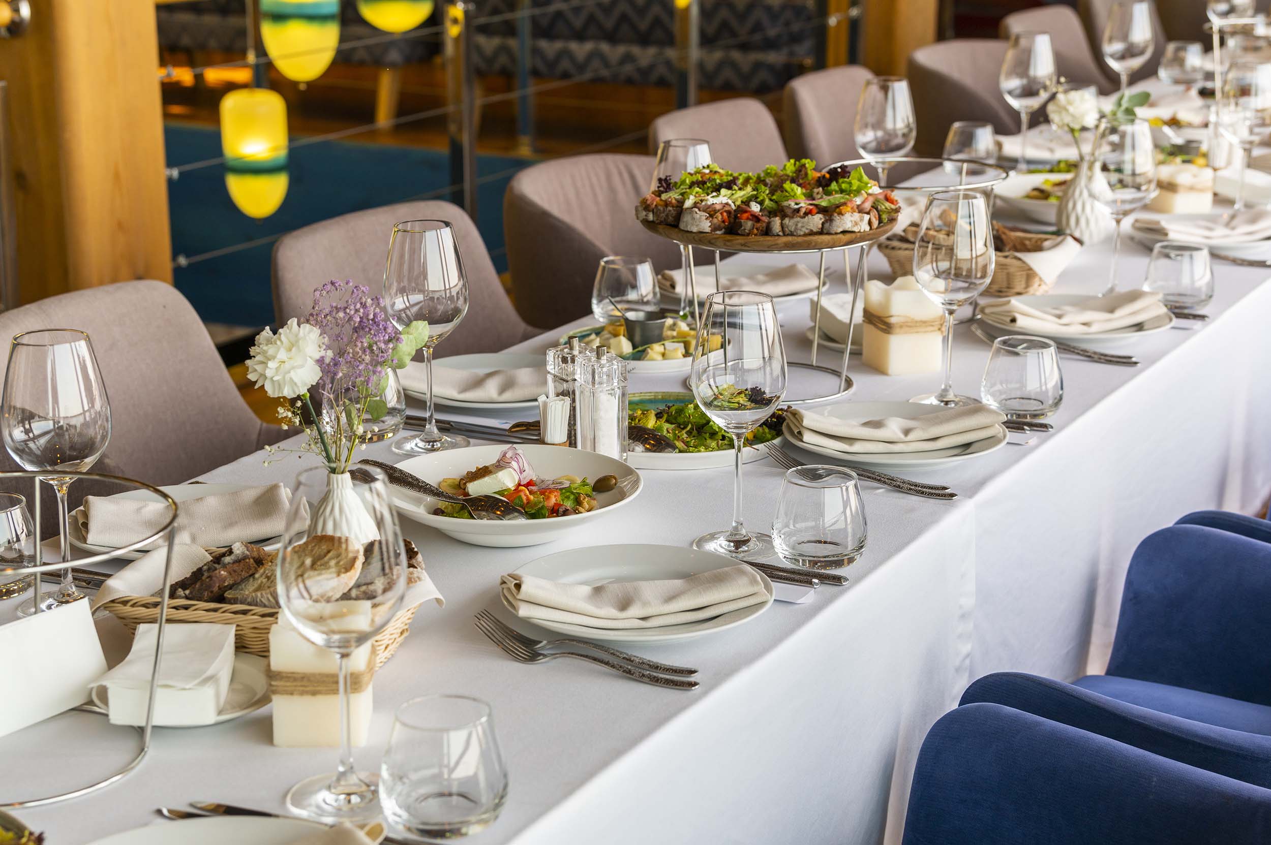 A long dining table is lavishly set with various dishes, glasses, and food, including salads and canapés, inside a well-lit restaurant with colorful yellow and blue accents.