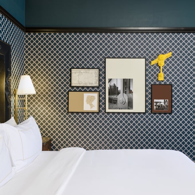 A king bed with white linens against a blue patterned wallpaper, flanked by a modern brass wall lamp.