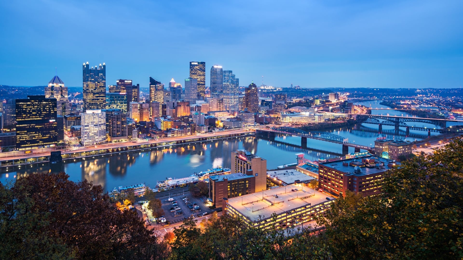 Panoramic view of the illuminated skyline at twilight, showing downtown buildings reflected in the river with several bridges spanning the water.