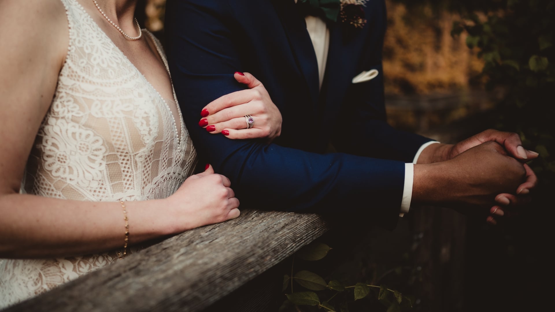 Close-up of a bride in a lace dress and a groom in a navy suit intimately holding arms and leaning on a rustic wooden rail outdoors.