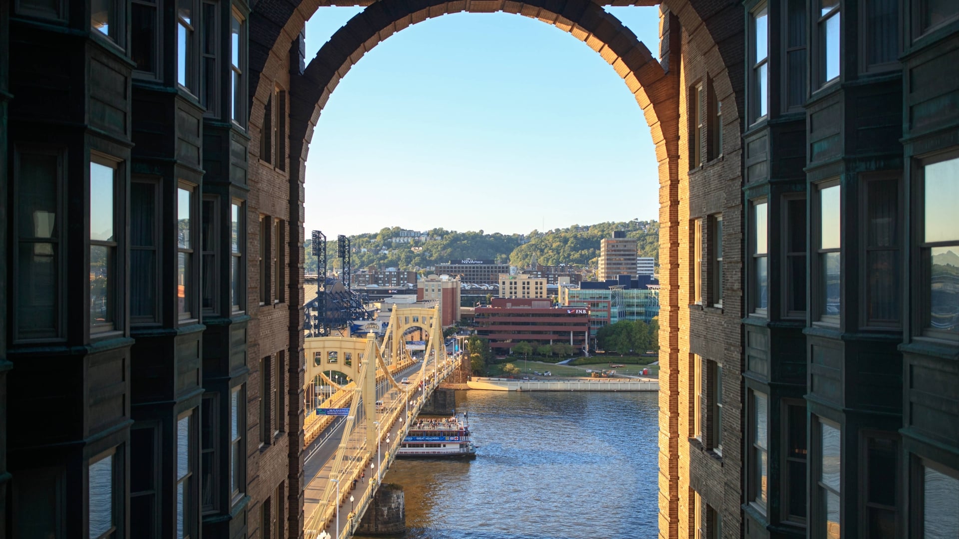 View of a yellow suspension bridge and the Pittsburgh skyline, framed through a large stone archway.