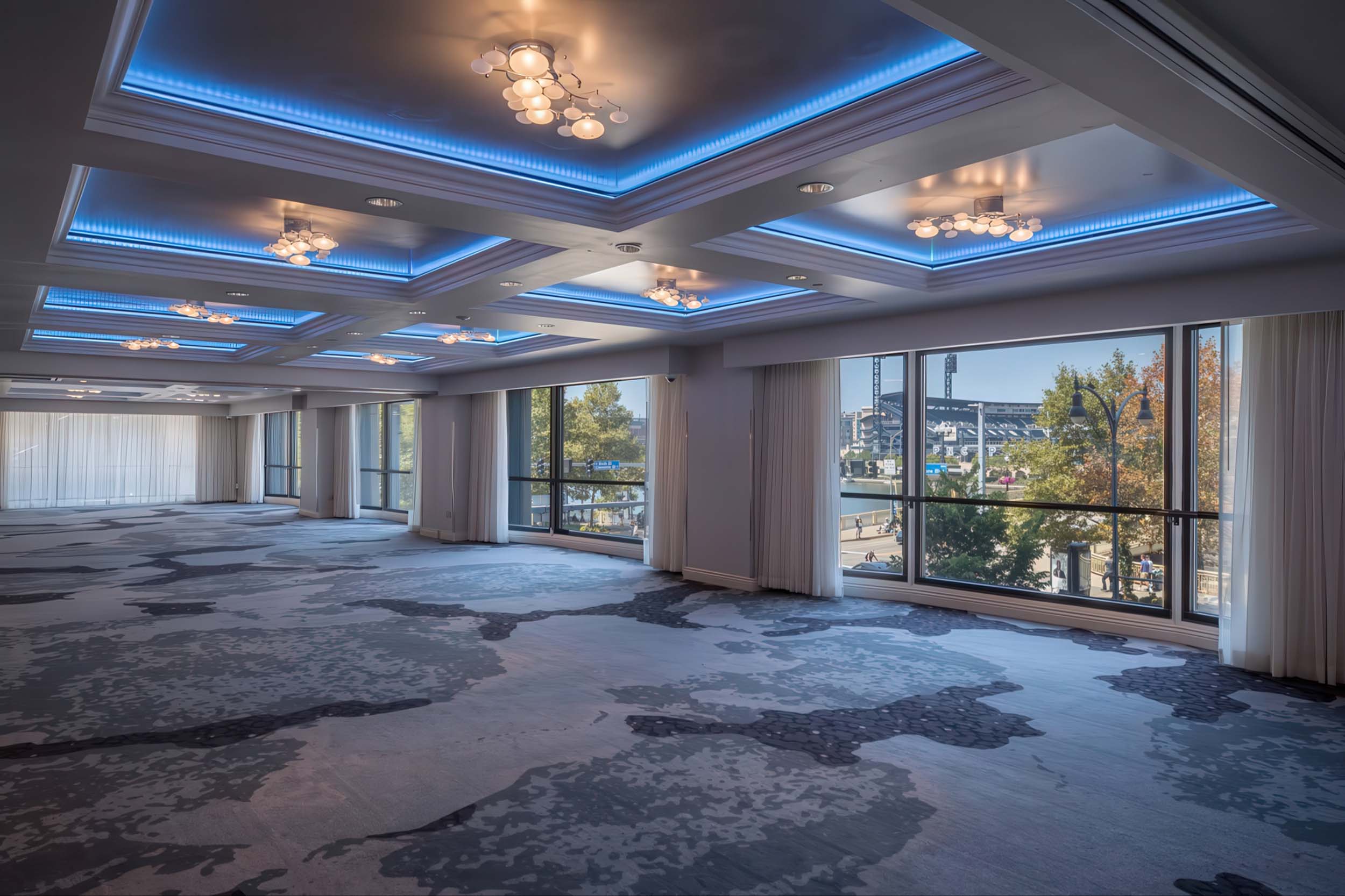 Large, empty ballroom features a patterned carpet, modern ceiling chandeliers with blue LED lighting, and expansive windows overlooking an urban waterfront with PNC Park stadium.