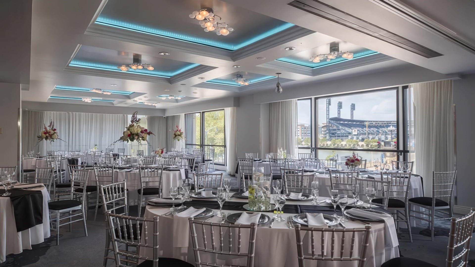Event room. Round tables are formally set with black and white linens and chairs, featuring centerpieces. Large windows reveal a baseball stadium and city view.
