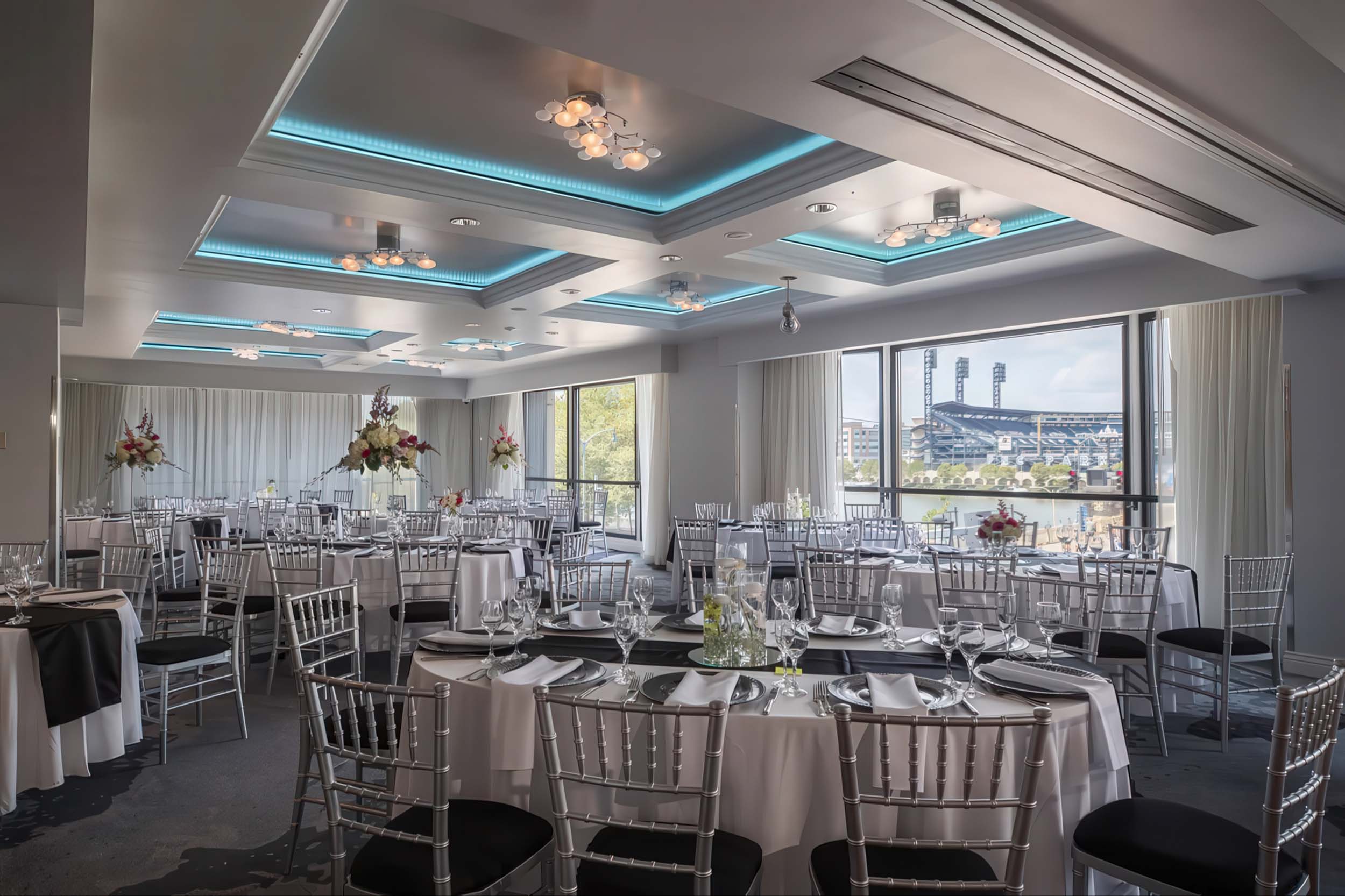 Event room. Round tables are formally set with black and white linens and chairs, featuring centerpieces. Large windows reveal a baseball stadium and city view.