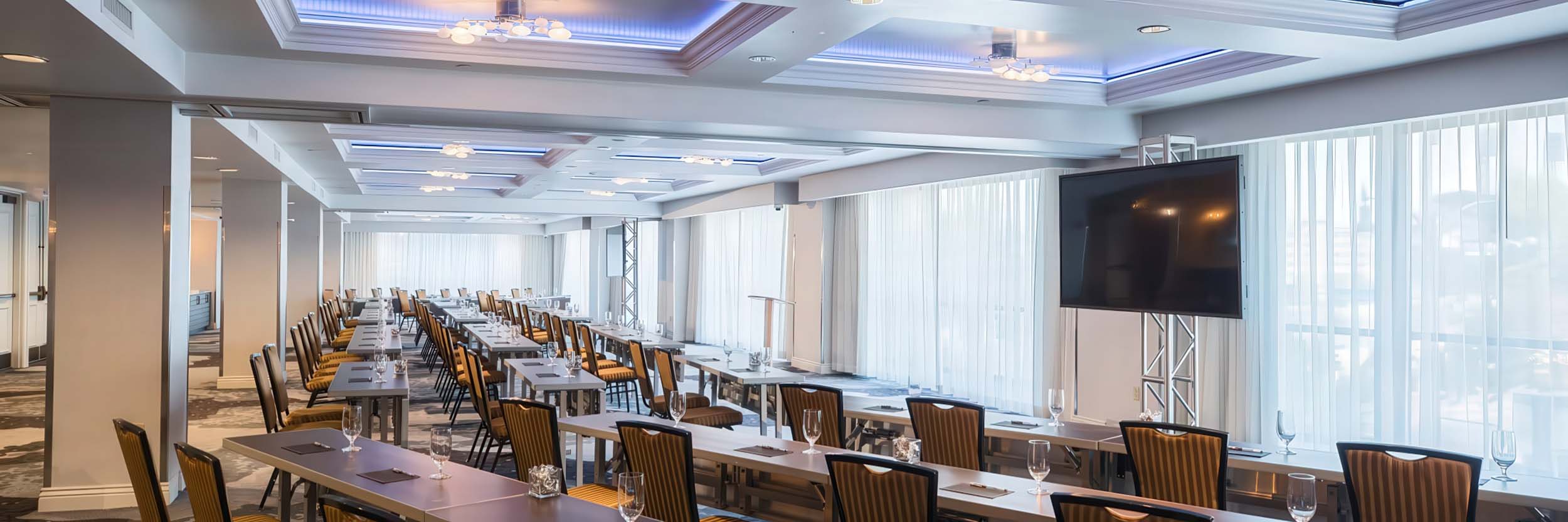 A spacious meeting room is prepared with multiple rows of tables and chairs, each set with glasses and notepads, facing a large TV screen, framed by sheer window curtains.