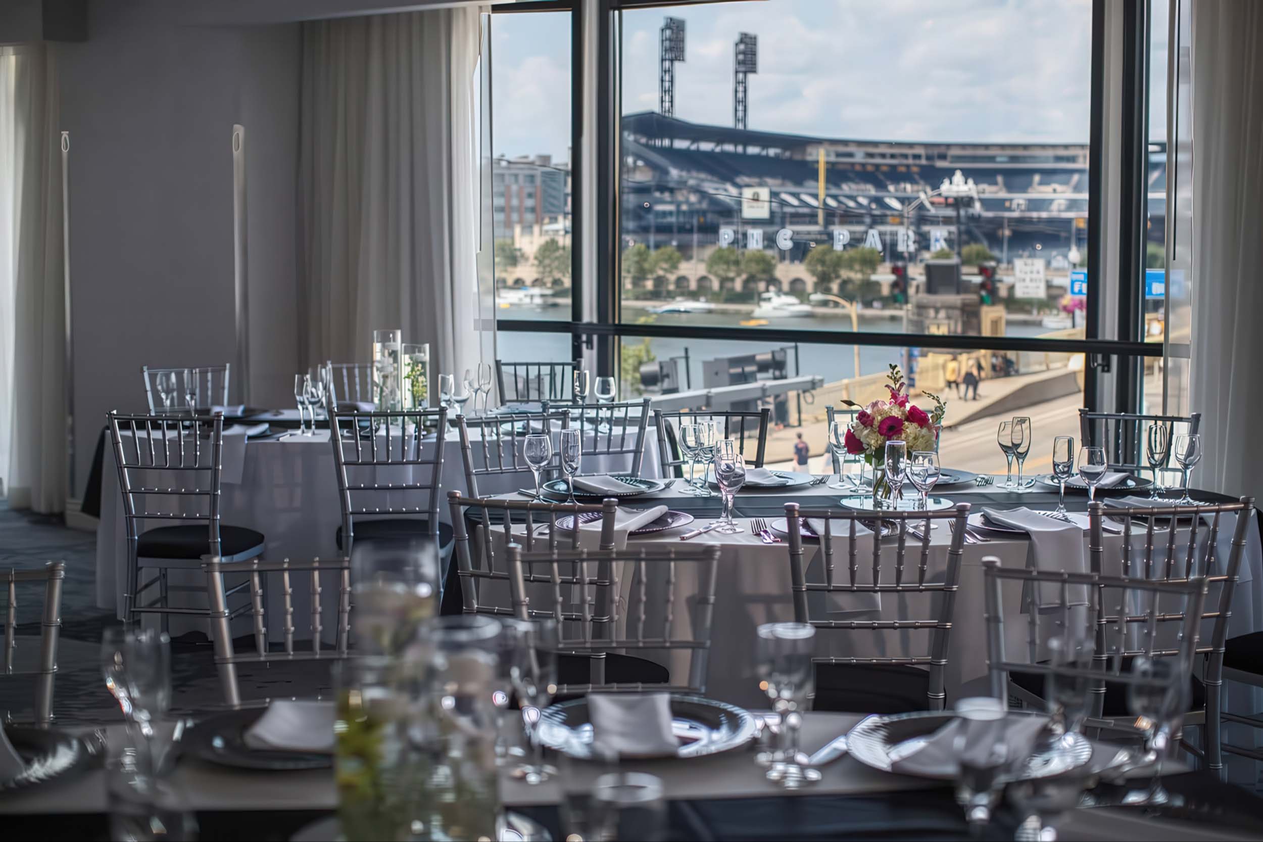 Elegant dining tables, set with place settings and floral arrangements, are arranged inside a room with large windows overlooking PNC PARK and a river with boats.
