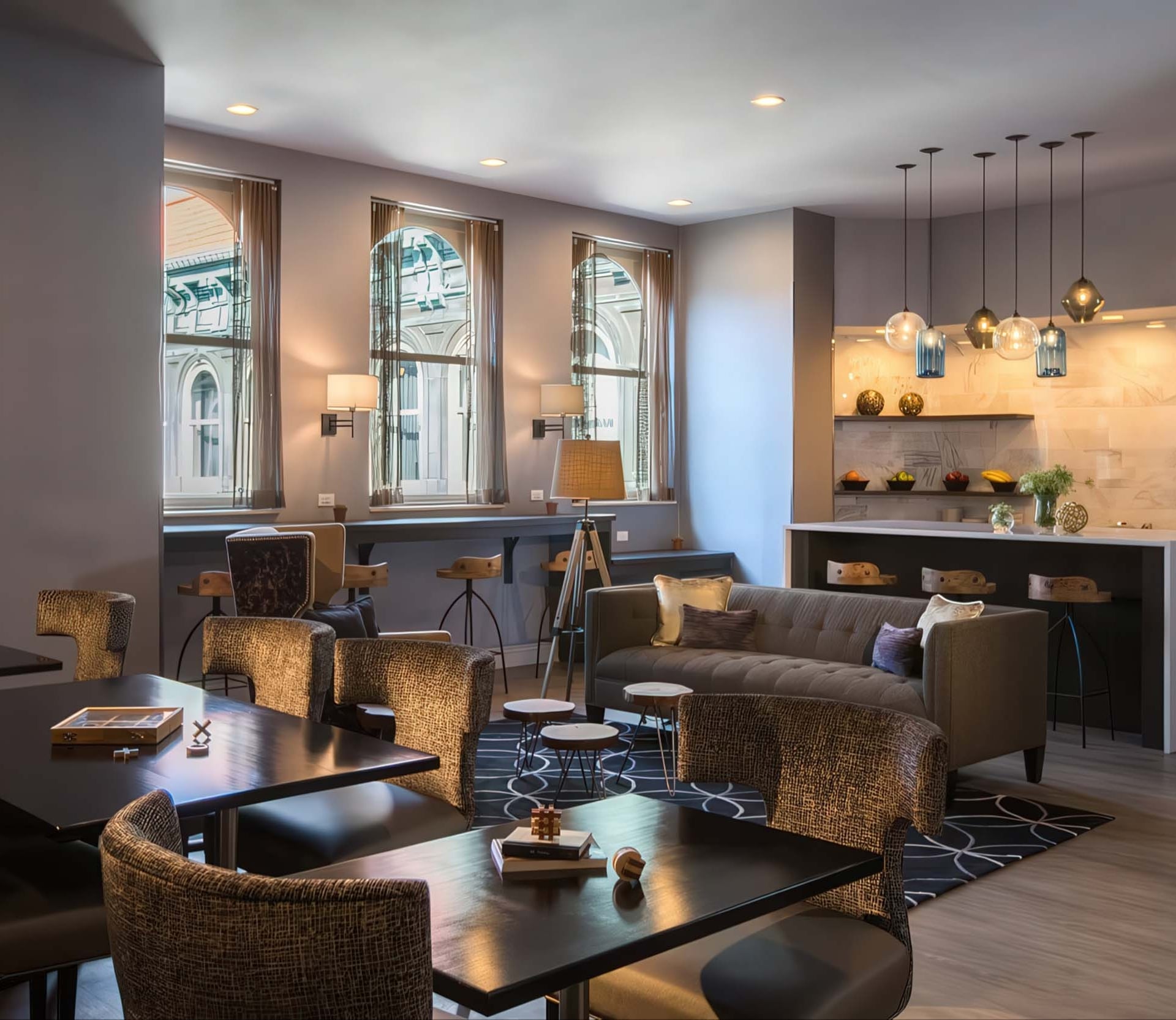 A modern lounge displays varied seating: dark tables with textured chairs, a grey sofa, and bar stools at a lit counter. Arched windows reveal an exterior building.
