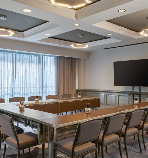 Conference room set up in a U-shape layout with modern chairs, a wall-mounted TV, and large curtained windows.