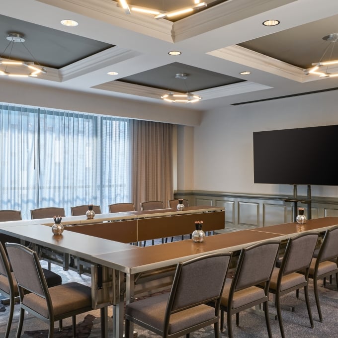 Conference room set up in a U-shape layout with modern chairs, a wall-mounted TV, and large curtained windows.