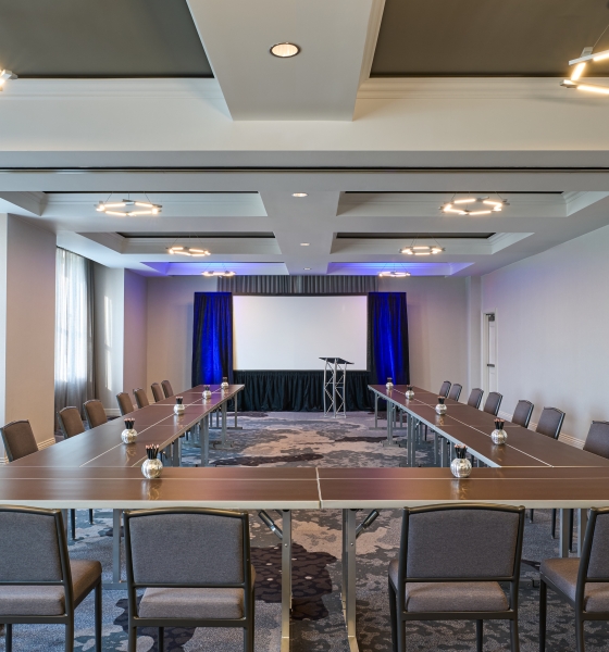 Conference room arranged in a U-shape with brown tables, gray chairs, and a presentation area with a white screen and blue drapes at the back.