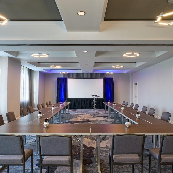 Conference room arranged in a U-shape with brown tables, gray chairs, and a presentation area with a white screen and blue drapes at the back.