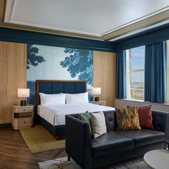 A modern hotel bedroom featuring a neatly made bed, leather sofa, and large windows revealing a city skyline with a baseball stadium in the distance.