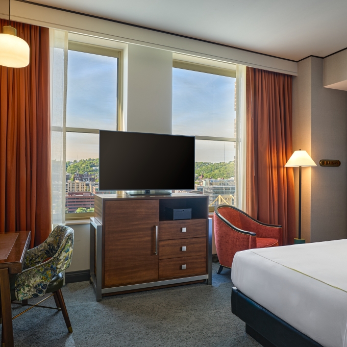 Stylish hotel room with a dark wood dresser, wall-mounted TV, desk setup, rust-colored curtains, and a plush bed.