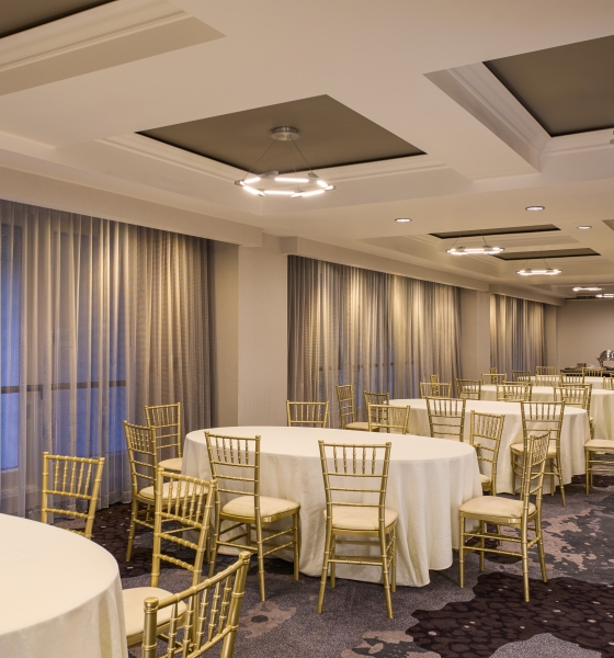Spacious hotel room setup featuring multiple neatly dressed round banquet tables with elegant gold chairs.