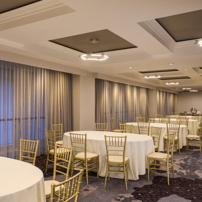 Spacious hotel room setup featuring multiple neatly dressed round banquet tables with elegant gold chairs.