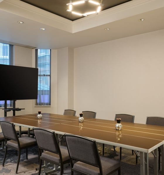 Conference room with a long brown table, modern gray chairs, a large TV display, and a geometric light fixture.