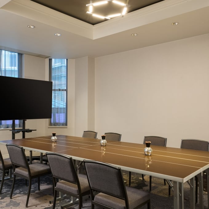 Conference room with a long brown table, modern gray chairs, a large TV display, and a geometric light fixture.