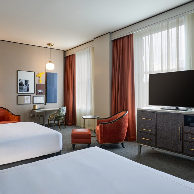 Modern hotel room with two queen beds, a flat-screen TV on a dresser, and large windows with orange curtains.