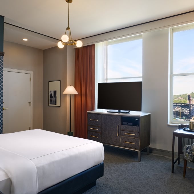 Modern hotel room with a king bed, a sleek desk setup, a TV dresser, and large windows with orange drapes.