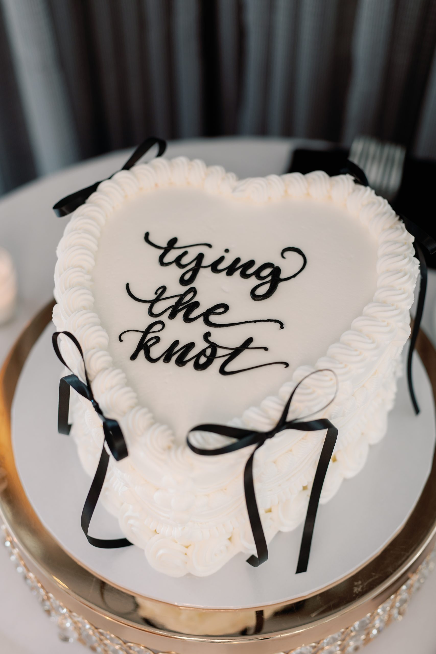 Wedding cake with white frosting, piped border, and black ribbon; top reads 'tying the knot'.