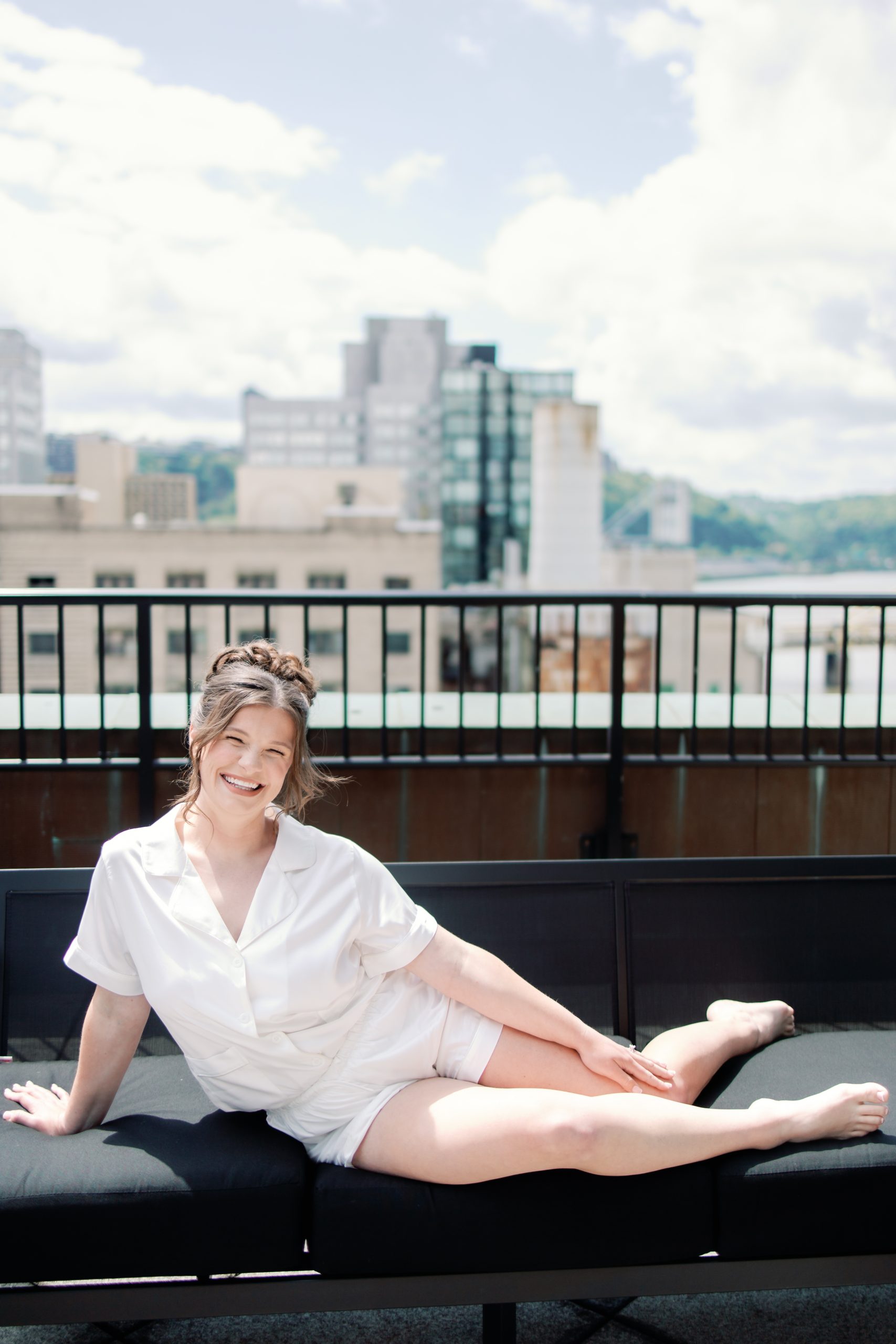 Smiling woman in a white blouse lounging on a black rooftop sofa with a city skyline behind the scene is visible in the background behind her.