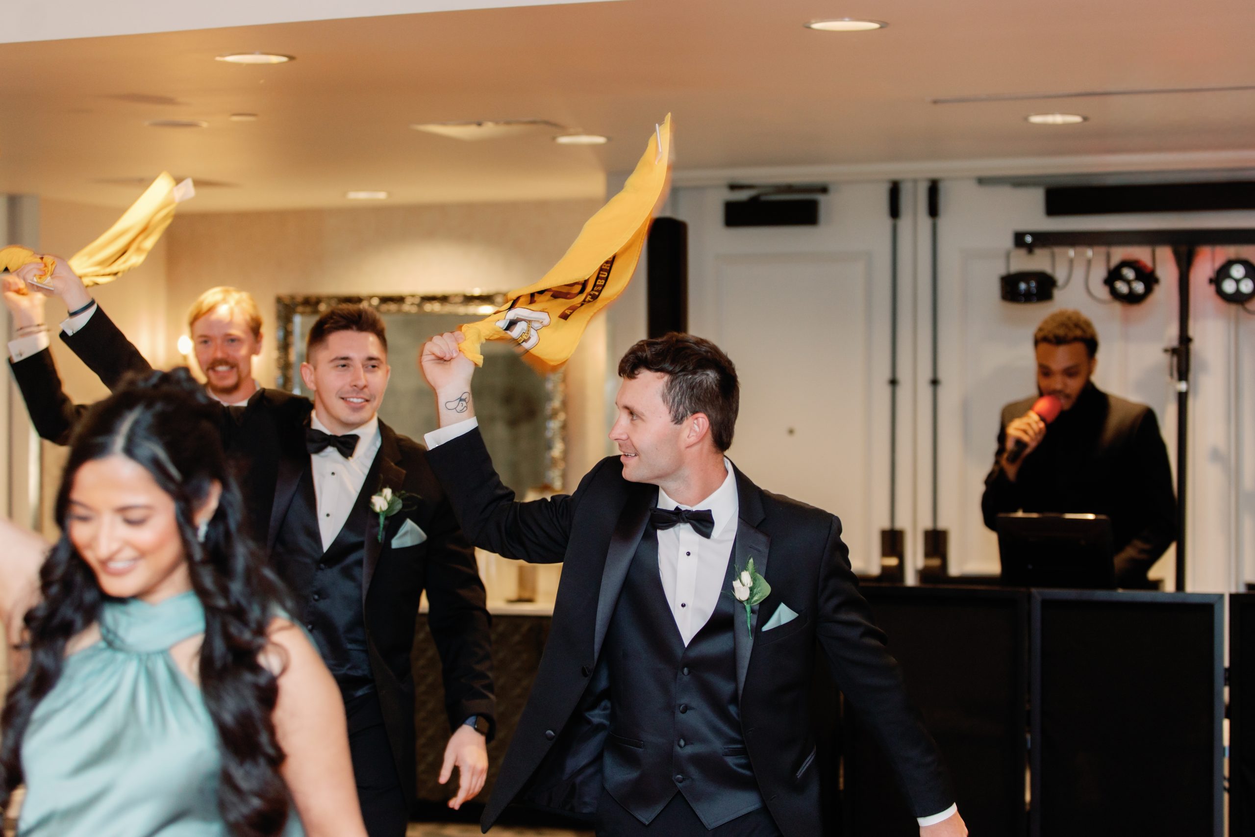 Men in tuxedos dancing and waving a yellow cloth at a wedding reception, with a singer in the background.