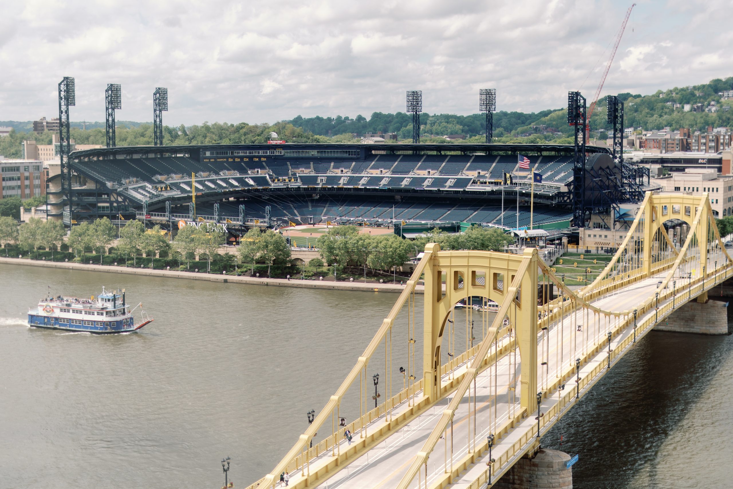 PNC Park on the riverfront with a yellow suspension bridge in the foreground and floodlit stadium seating behind it.