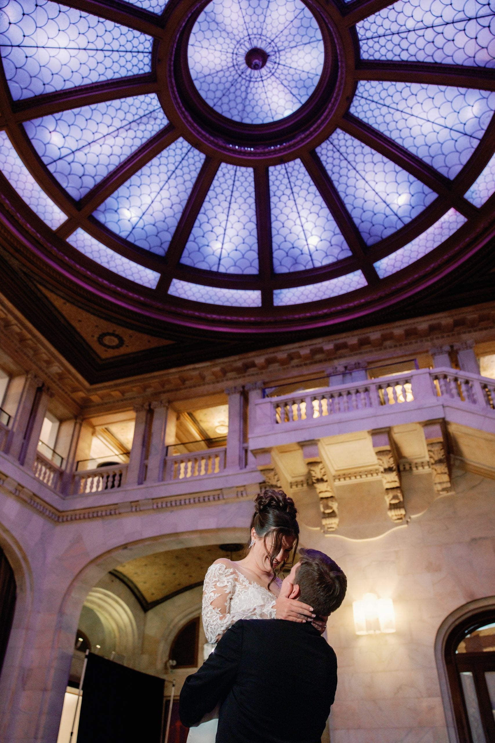 Couple kissing in a grand marble interior beneath a circular decorative skylight and balconies overhead.