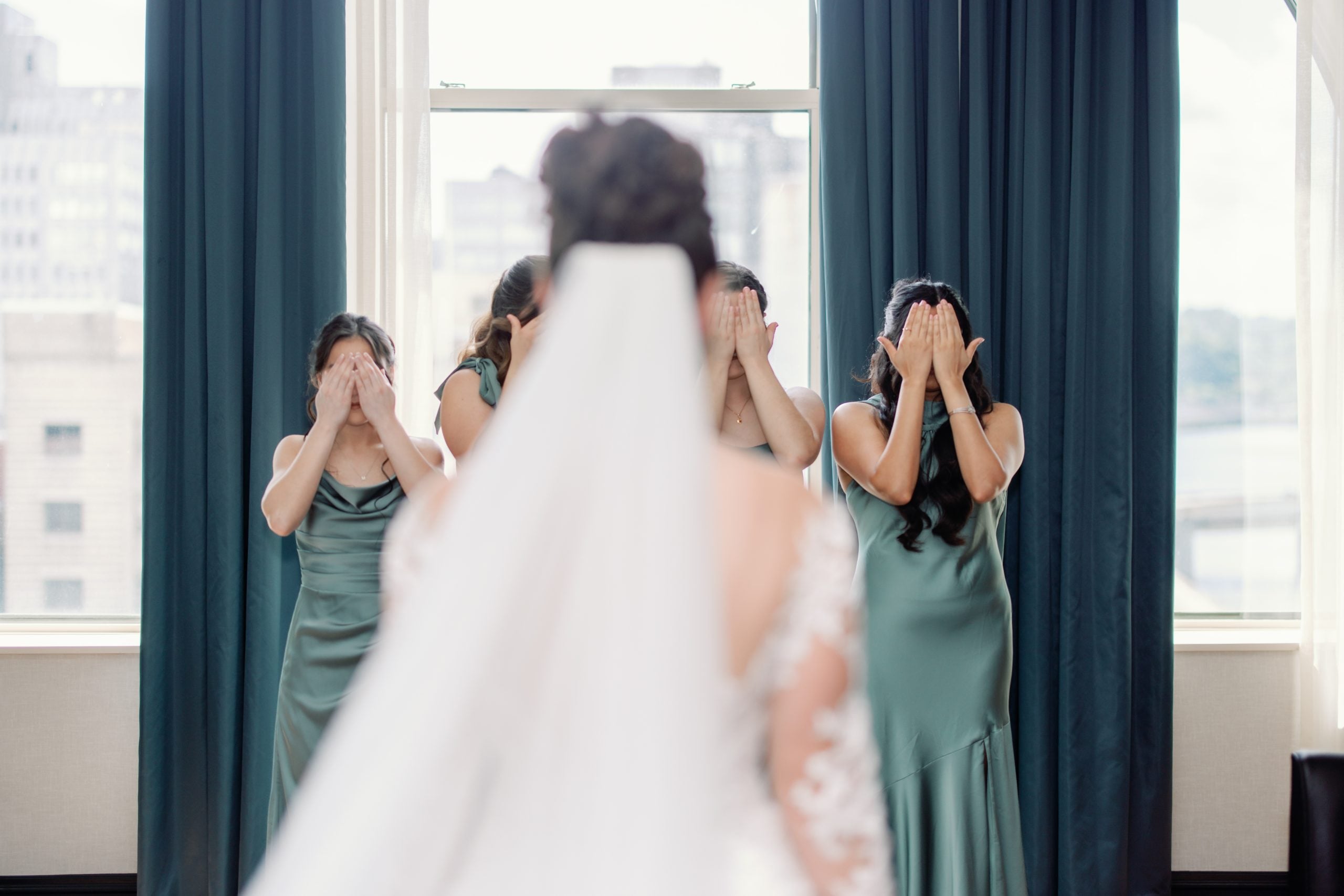 Bride in a white dress and veil passes by bridesmaids in green gowns who cover their faces with their hands by a window with blue curtains.