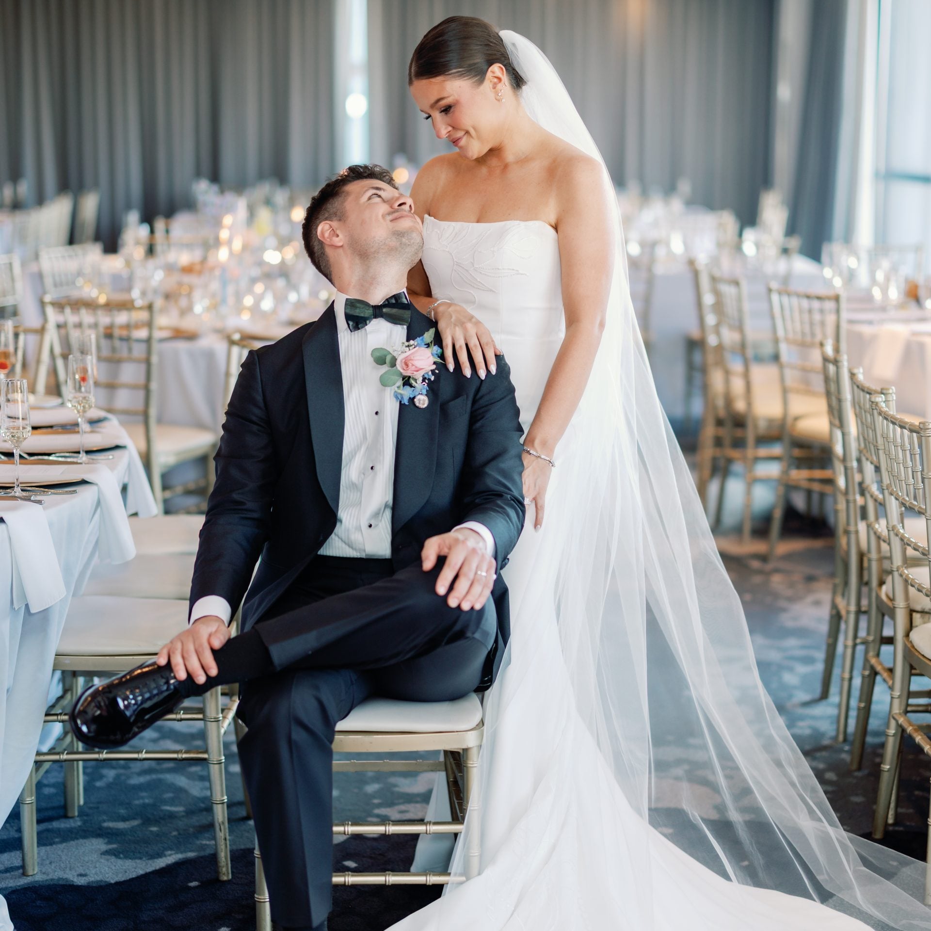 Bride in a strapless white gown and veil stands beside a seated groom in a black tux at a decorated wedding reception venue.