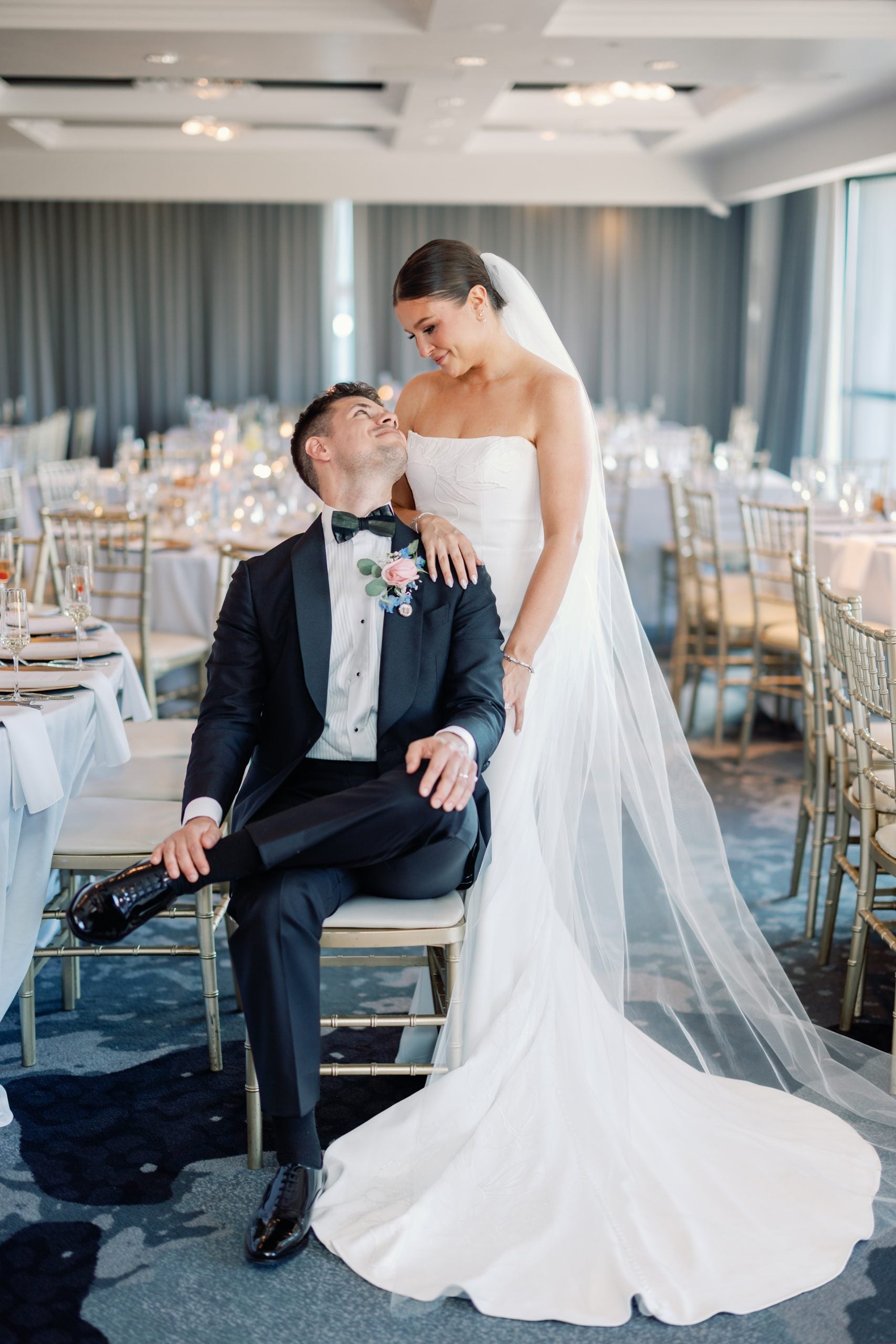 Bride in a strapless white gown and veil stands beside a seated groom in a black tux at a decorated wedding reception venue.