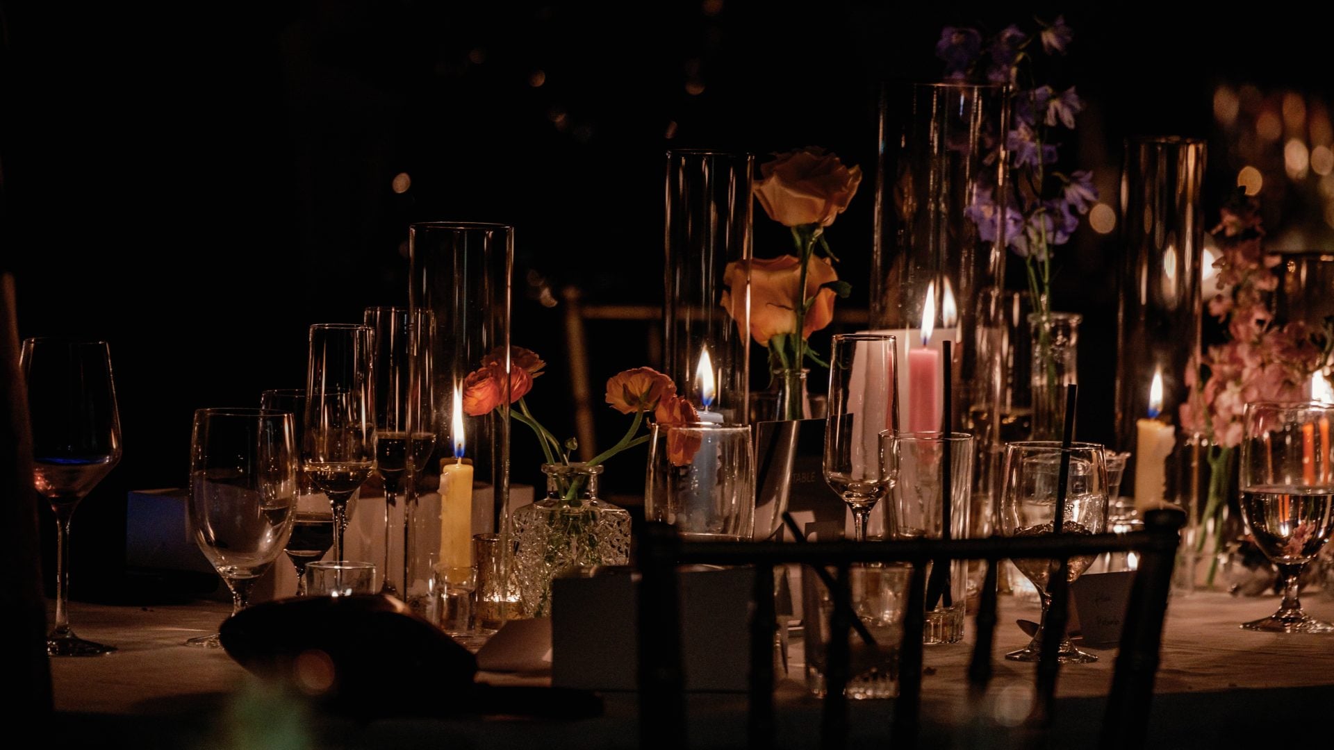 Dimly lit banquet table with candles, glassware, and floral centerpieces in a formal dinner setting at night.