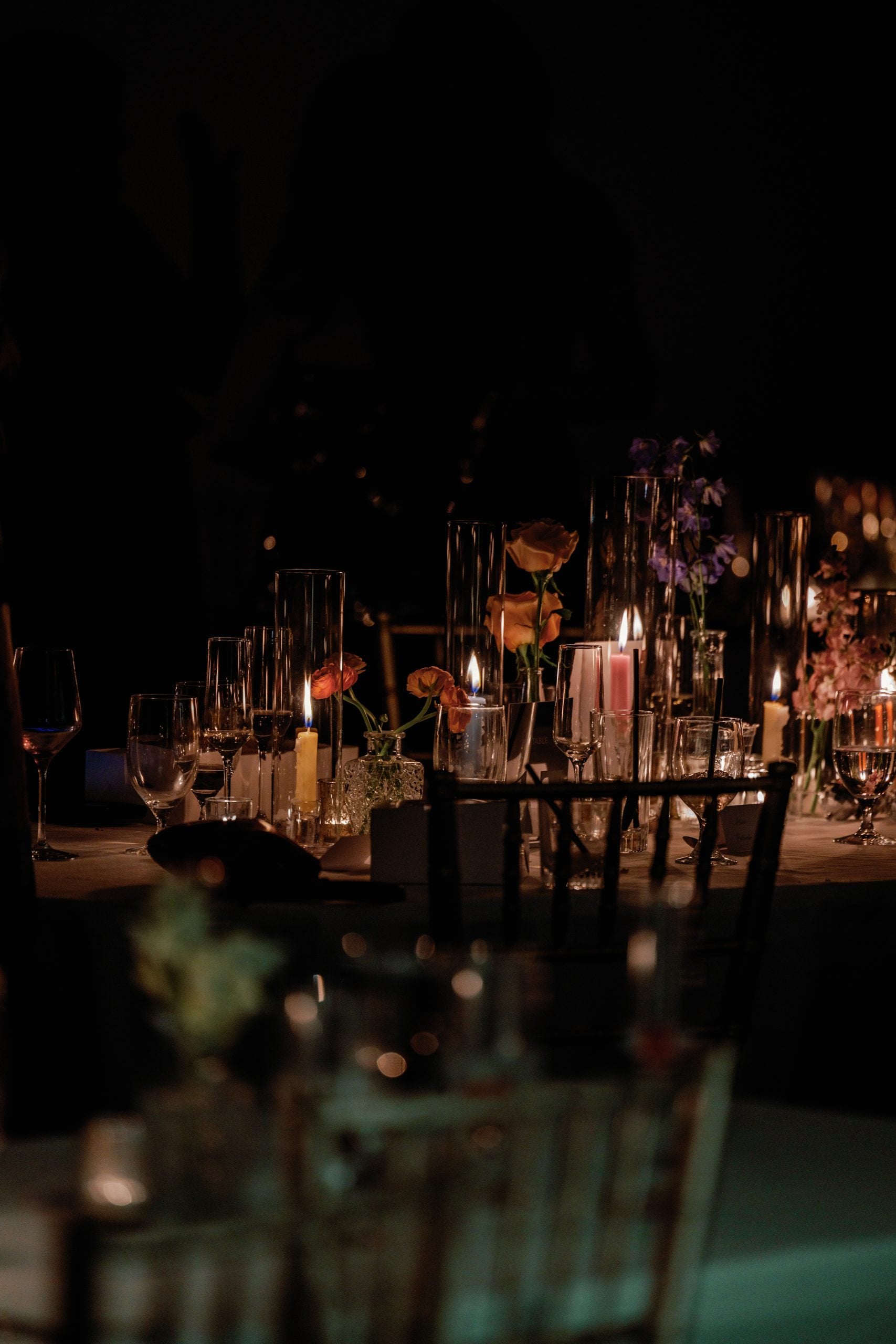 Dimly lit banquet table with candles, glassware, and floral centerpieces in a formal dinner setting at night.