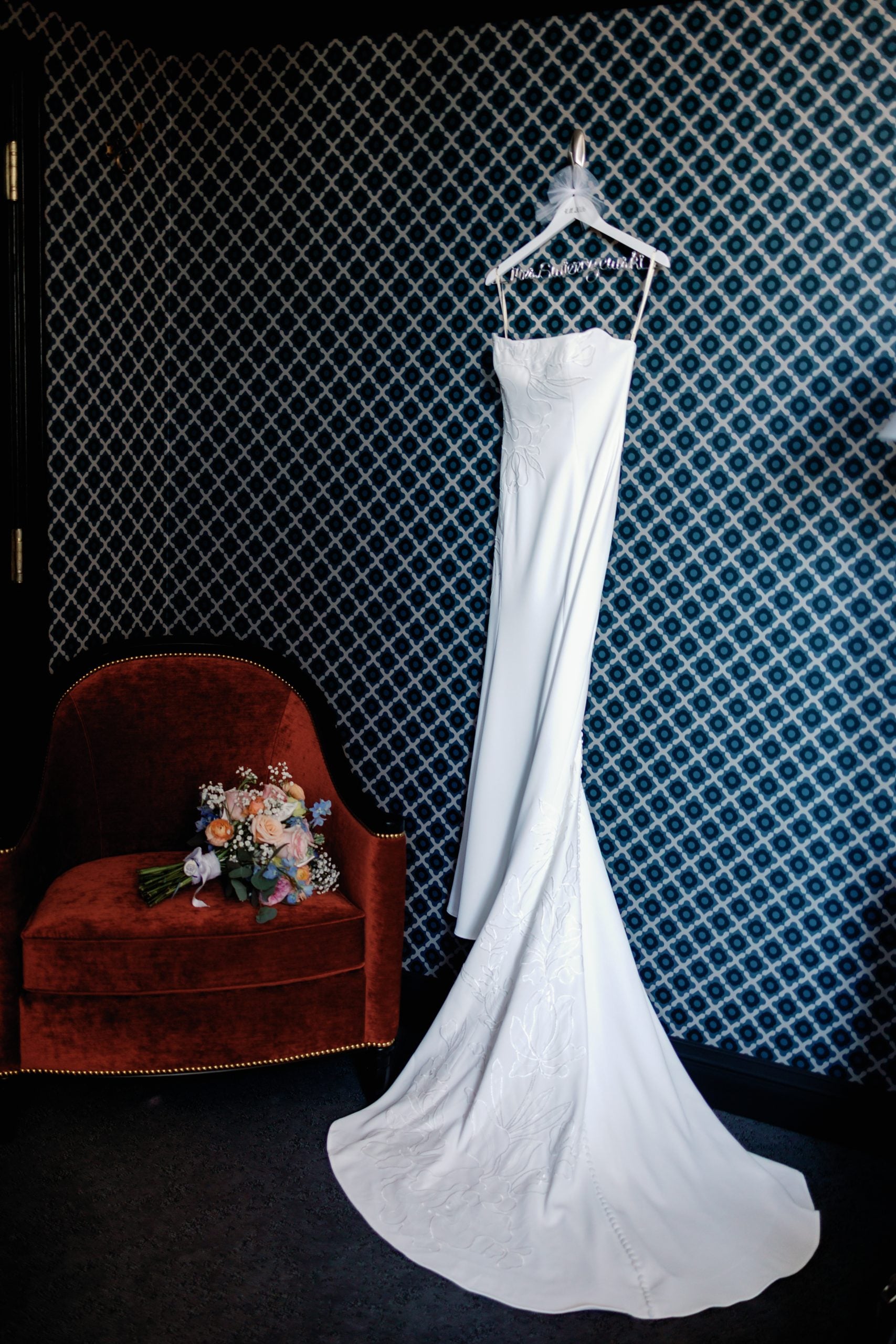 White wedding dress on a hanger against blue patterned wallpaper, with a bouquet on a red velvet chair nearby.