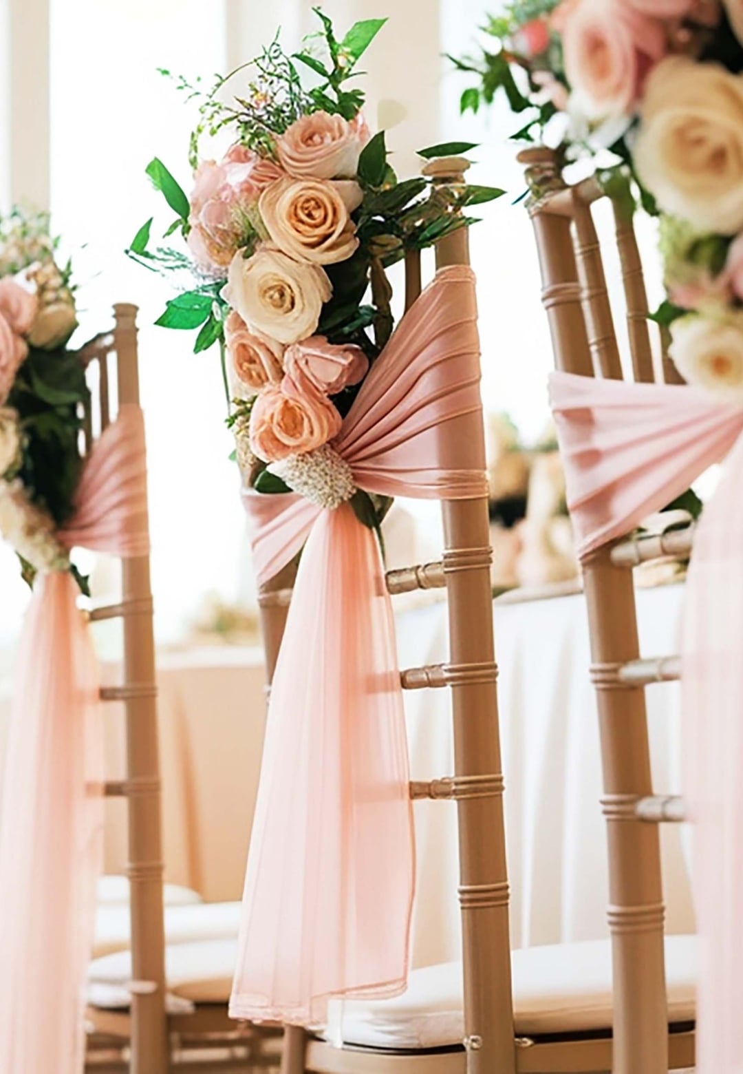 Row of wedding chairs adorned with blush pink roses, greenery, and pink fabric sashes along an aisle.