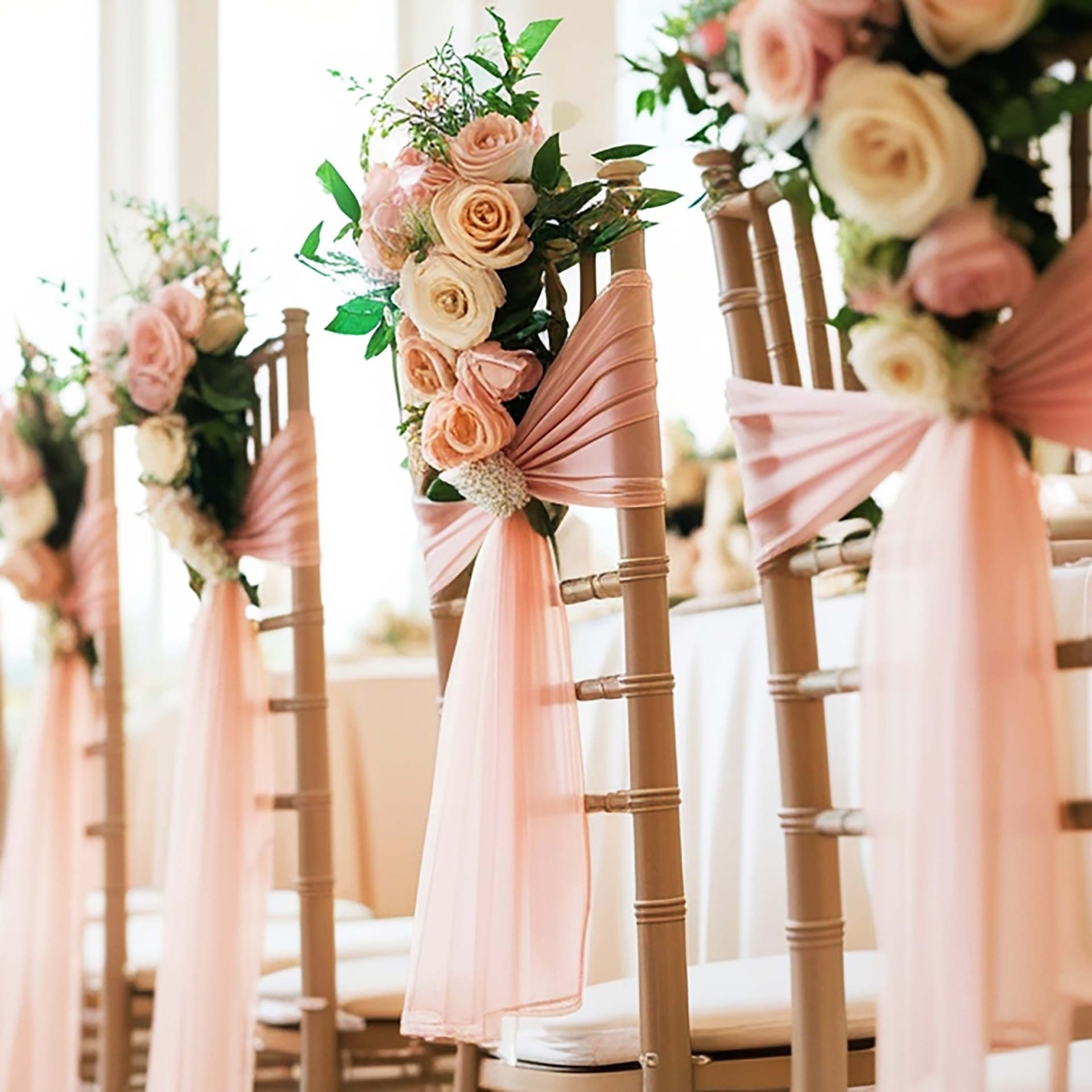 Row of wedding chairs adorned with blush pink roses, greenery, and pink fabric sashes along an aisle.