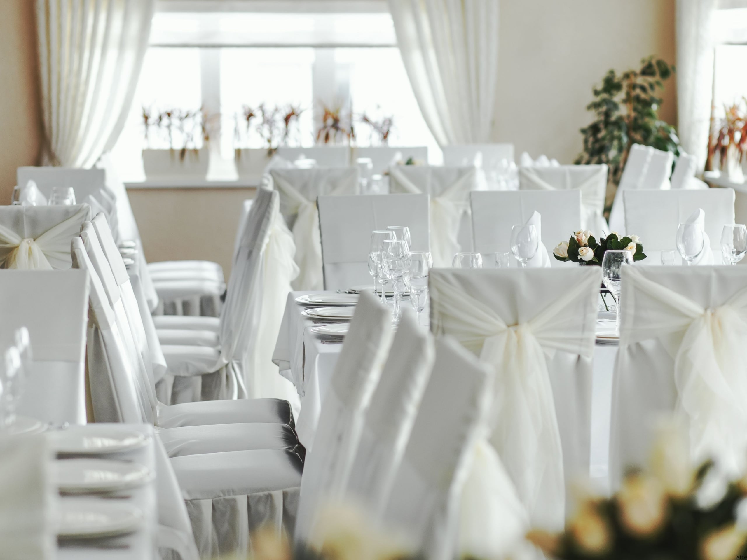 White-draped banquet tables set with glassware in a sunlit, elegant reception hall.